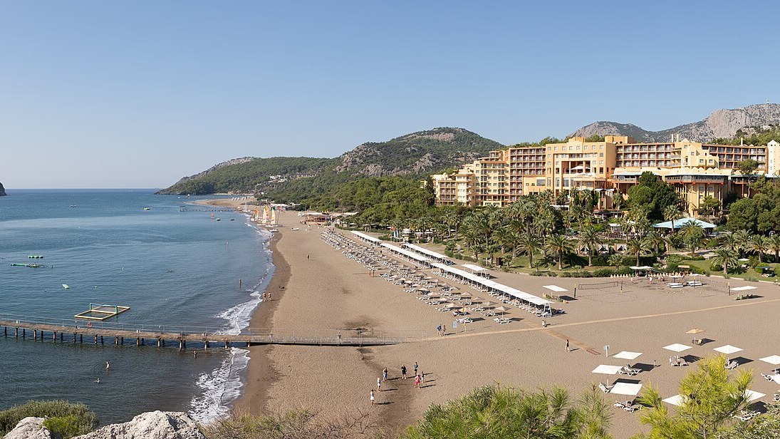 Sarıgerme kıyısında otel binaları, kumsal boyunca şezlonglar ve arkada Baba Adası